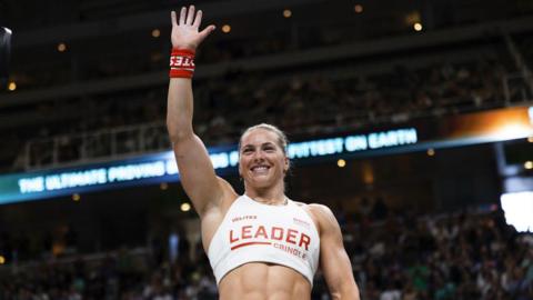 Cringle waiving to the crowd in the arena. She is wearing a white vest with leader on the front in red lettering and her name beneath it.