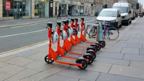 Six orange e-scooters are lined up on the pavement in Grainger Street, Newcastle. Shops, including a sports shop, are on the other side of the road.