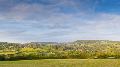 A wide vista of a typical Cotswold landscape of rolling hills and woodland copses.  A big blue sky with whispy cirrus clouds. 