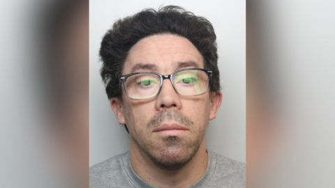 Shaun Smith has medium length slicked-back black hair, dark stubble and glasses.