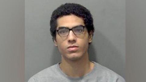Nicholas Prosper, who has dark hair and dark glasses, staring into the camera for a police custody image. He is wearing a grey sweatshirt.