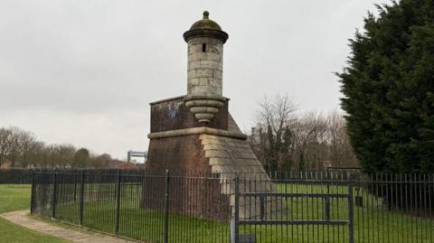 A 16th Century round stone watch tower with a small window and a spherical roof. The tower is mounted on a brick and stone plinth, which has been built to resemble part of a Tudor fortress, It is surrounded by metal fence and is part of a grassed park area, with green trees to the right.