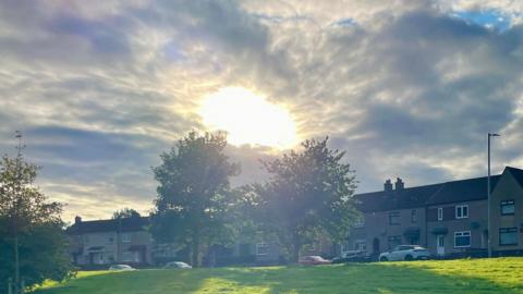 The Sun shines through a large circular gap in the clouds over trees and a row of houses