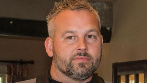 Daniel Johnson has salt-and-pepper hair that is slightly longer on the top and shorter on the sides. He has a darker beard and is slightly looking off to the side of the camera.