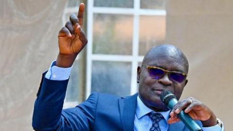 Simon Byabakama in glasses, wearing a blue suit and a dotted tie and speaking into a mic 