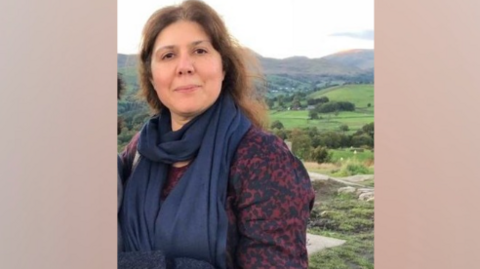 Dr Shaista Shirazi is standing and directly facing the camera with a slight smile on her face. The rolling hills of the countryside are behind her. She has long brown hair and is wearing a blue scarf and purple top. 