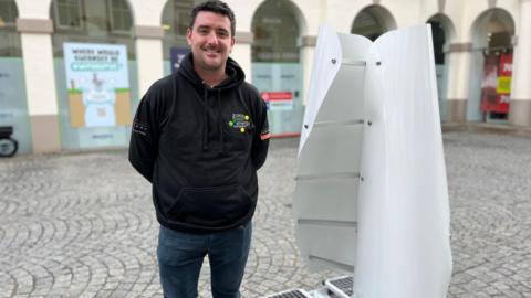 A man stands next to a white wind turbine, which is made of two curved, vertical sails on a flat base. The turbine is slightly shorter than the man.