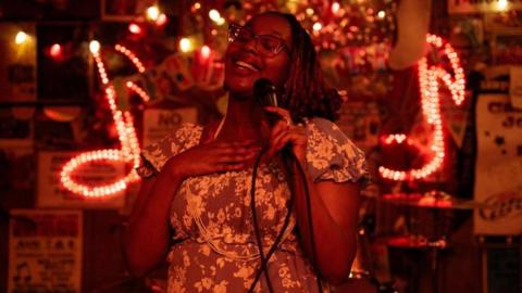 Edna Nicole Luckett sings at a microphone, her hand on her heart, with light-up signs showing musical notes behind her