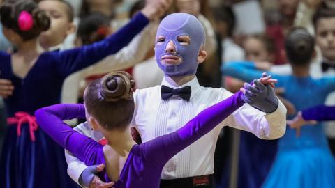 Roman Oleksiv, wearing a purple burns mask, white shirt and bow tie, dances with girl wearing purple. Many other dancing couples are behind them