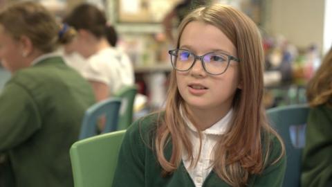 Girl in green school uniform with long brown hair and glasses sitting in a classroom with other children in the background.