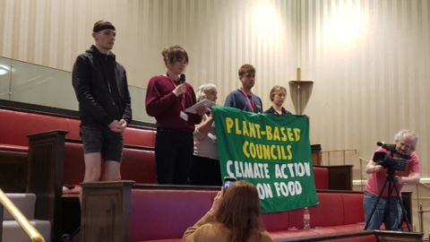 Image shows five vegan campaigners standing in the stalls at a council meeting, with three campaigners holding a green banner that reads 'PLANT-BASED COUNCILS CLIMATE ACTION ON FOOD' 