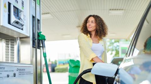 A woman with short curly brown hair with blonde highlights filling her car up at a petrol station.