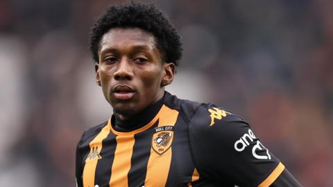 A head and shoulders picture of Jaden Philogene, wearing Hull's black and gold striped top, as he looks across the camera to something out of sight.