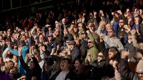 A packed scene of fans standing up, with arms raised, in the stadium.
