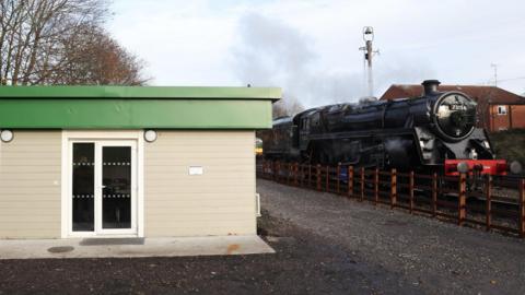 A black steam engine parked next to a cream building with a green roof