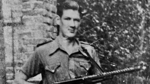 A black and white photograph shows a young man with slicked back dark hair looking at the camera. He is wearing a short-sleeved military shirt and carrying a rifle.