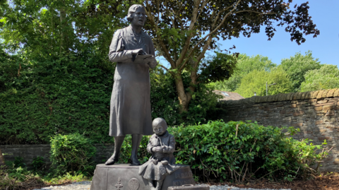 A grey statue of a woman, stood on a plynth, with a statue of a child sat down at her feet, clutching a teddy bear. 
