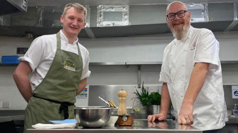 Two chefs stood in the center of a kitchen either side of a workspace. The man on the left, Harrison, is stood wearing a green apron with his hands behind his back. The man on the right, Simon, is wearing his chefs whites and is leaning on the workbench.