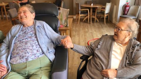 Ruth Bester and Brenda Sired sitting in chairs and smiling as they hold hands