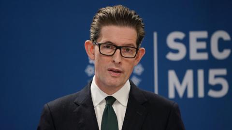 Director General of MI5 Ken McCallum, a man with black-rimmed glasses and swept-back dark hair, wearing a dark suit and tie, standing in front a navy blue backdrop with the MI5/Security Service logo