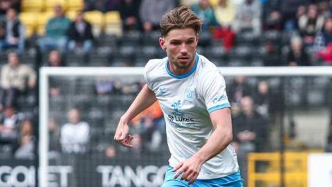 Joel Tabiner, in a white Crewe away top, looks down at the ball during a League Two game before his injury