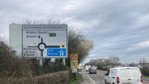 Cars driving towards a roundabout. There is a sign on the left pointing drivers in the direction of the M5 and the Bristol Airport.