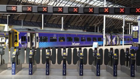 A line of ticket barriers at a railway station. The signs overhead all have red crosses. A blue train can be seen waiting at the platform behind the barriers.