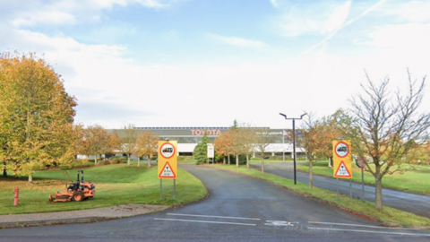 Two yellow signs in front of a patch of grass, which is in front of the Toyota manufacturing plant