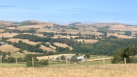 The view from Nantglyn, Denbigshire of an agricultural landscape covered with parched fields. There is a fence in the foreground, with dry hills in the background.