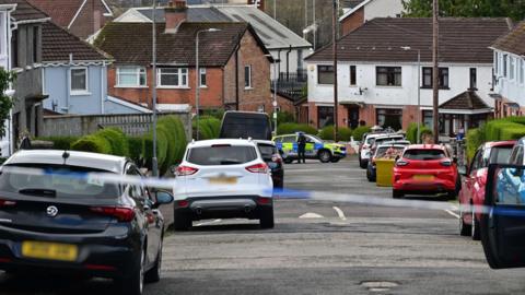 A row of multiple cars sit either side of a residential road. There are red-brick houses the run the length of the road. At the bottom of the road a blue and yellow police car sits behind blue and white police tape. A police officer in black uniform stands beside the car. 