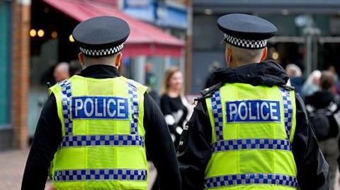 The backs of two police officers who are walking away from the camera. They are patrolling a high street and are both wearing black police uniform caps, black jackets and a yellow fluorescent vest which has the word POLICE on the back in blue and white writing.