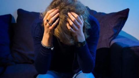 A woman sits on a dark blue sofa with her head in her hands. She is wearing rings and a necklace, a navy blue jumper and light blue jeans.