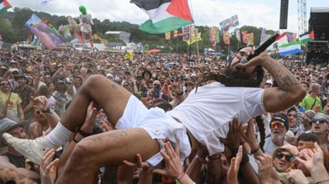 A photograph of Bob Vylan crowdsurfing during his Glastonbury set. He appears to be floating on top of the crowd as they hold him up using their hands. He is wearing a white t-shirt and white shorts. 