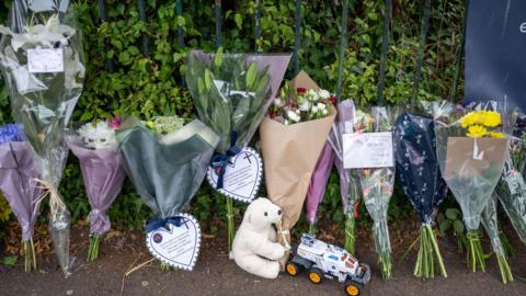 Several bouquets of flowers laid in tribute to a boy who died. Many of them include notes and cards. There is also a teddy and a toy car on the ground.
