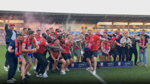 A football team in red shirts lift a trophy and spray champagne