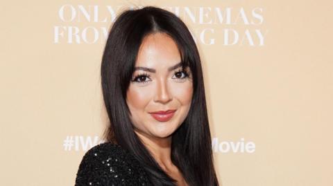 Sharon Gaffka has long dark hair and is wearing a black sequined dress. She is standing in front of an ivory wall.
