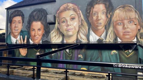 The Derry Girls mural in Derry, featuring four of the characters from the TV show. They are dressed in school uniforms. In the foreground is a seating area while an awning that partially covers the mural is being reinstalled