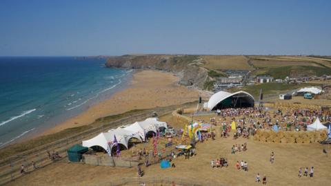 A large outdoor festival near a beach, featuring white tents, colorful structures, and crowds of people on a grassy area. A stage with a white canopy is visible in the background, while attendees enjoy the beach along a scenic coastline with cliffs and hills.