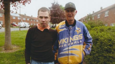Two men standing on a suburban pavement, one wearing a black sweater and the other a blue and yellow Vikings American Football jacket.