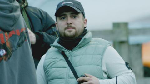 Connor Graham standing behind a metal barrier at a protest. He has a beard and is wearing a black baseball cap, light grey top and a bodywarmer.
