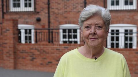 A woman with short light-grey hair stands in front of a large red-brick building. She is wearing a yellow jumper, gold ear-rings and has a serious expression on her face.