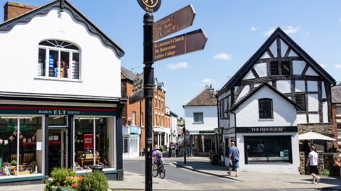 A number of black and white timbered shops on a narrow street with people walking and cycling on it