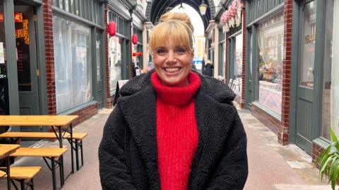 Steph Ellis pictured standing inside the arcade, She has light-coloured hair and is wearing a black jacket and red jumper. She is smiling at the camera. There are shops on either side in the background.
