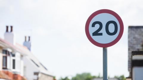 Red 20mph road sign with homes in the background.
