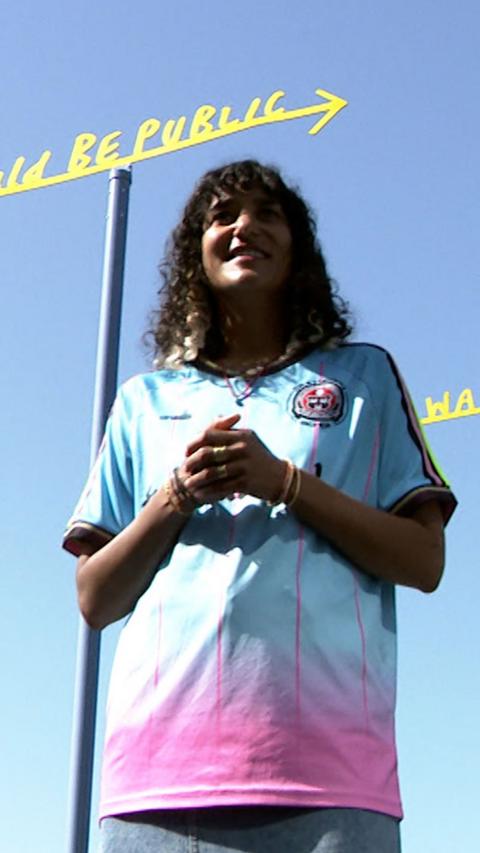 A smiling woman in a blue and pink top stands in front of a tall blue structure with gold lettering at the top.
