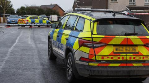 A still of two marked police cars parked in a housing estate. In the distance is some red and white, as well as blue and white police tape cordoning off the area.