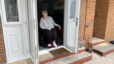 A woman in her sixties is sitting in a wheelchair inside a house looking out of some open patio doors.  Leading up to the doors are two steps.