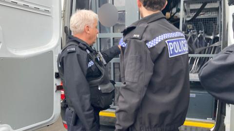 Two police officers are shutting the door to a police van, as someone who has been arrested sits inside