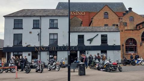 King Billy Rock Bar, a light blue and black building with three floors on the left side and two storeys on the right. Outside are several motorbikes and motorbike riders. In the background was a red brick casino.