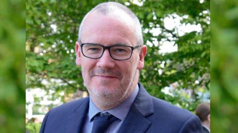 Kevin Flanagan, with short grey hair and dark framed glasses, wears a navy suit jacket, blue shirt and navy tie.